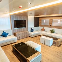 Moderne yacht lounge med hvid hjørnesofa, farverige puder, trægulv, væg-TV og stemningsbelysning i loftet.