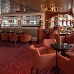 Elegant salon ombord på skibet Switzerland med koralfarvet lænestole, runde borde, rødt tæppe og træpaneleret loft med lysekroner.