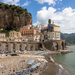 Kystby Atrani på Amalfikysten med historiske bygninger i klipper, buekonstruktioner og strand med parasoller og både.