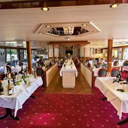 Elegant restaurantinteriør på MS Olympia med hvide duge, bordeauxrøde stole, rødt tæppe og store vinduer med udsigt over vandet.