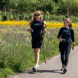 Two women walking on paved path with hiking poles and backpacks. Wildflowers and yellow blooms line the path, with vineyard posts visible in background.