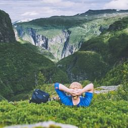 Vandrer i blå jakke hviler på græs med hænderne bag hovedet og ser ud over grøn bjergdal i Aurlandsdalen, Norge.