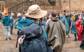 Gruppe Camino de Santiago pilgrimme med rygsække og vandrestave forsamlet på en stengård, iført udendørs udstyr og solhatte.