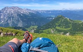 Vandrers ben med orange sko og blå rygsæk på alpeng, med udsigt over Watzmann og grønne dale i Berchtesgaden.