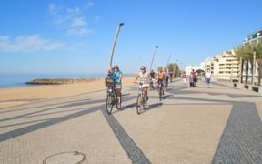 Grupp cyklister med cyklar på stenlagd kustpromenad i Algarve, Portugal. Strand, palmer och vita byggnader i bakgrunden.