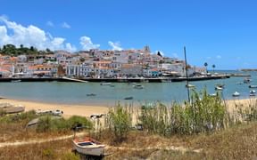 Pittoresk kuststad i Algarve med vita byggnader, sandstrand, turkos vik med båtar och liten fiskebåt i förgrunden.