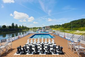 Soldäck på MS SE-MANON med jätteschackspel på schackrutigt golv, pool, trädäck och flodvy med gröna kullar.