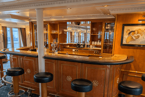 Elegant bar ombord på MS Casanova med buet trædisk, sorte lædertaburetter, udsmykket loft og vinduer med udsigt over vandet.