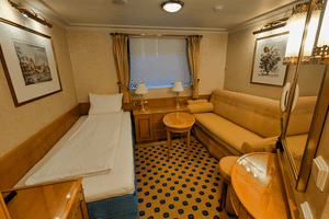 Kompakt kahyt på MS Casanova med enkelseng, beige sofa, rundt bord, vindue med gardiner og blåt mønstret tæppe.