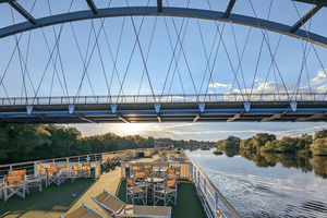 Udsigt fra MS Casanovas soldæk under passage af en moderne skråstagsbro. Liggestole og borde synlige på dækket med flod og træer forude.