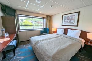 Modern skeppshytt på MS SE-MANON med dubbelsäng, stort fönster mot flod, skrivbord, fåtölj och inramad konst på väggen.