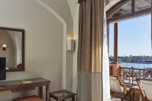 Lyst hotelværelse på Sultan Bey El Gouna med trævmøbler, TV, spejl og altan med trævstole med udsigt over marinaen.