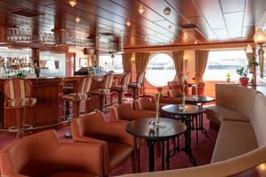 Elegant salonbar på skibet Switzerland med træbar, læderstole, runde borde og runde koøjer med udsigt over vandet.