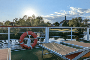 Soldæk på MS Casanova med rød redningskrans, liggestole og hvidt rækværk med udsigt over stille vand. Kirketårn og træer synlige i baggrunden.