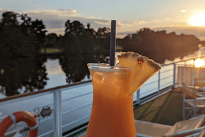 Orange tropisk drink med ananas pynt på MS Casanovas soldæks rækværk ved solnedgang, med rolig flod og træer i baggrunden.