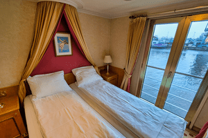 Kahyt med to senge på MS Casanova med hvidt sengetøj, gyldne gardiner, rød accentvæg og store vinduer med udsigt over havnen.