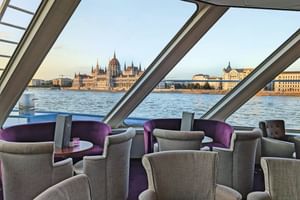 Interiör av fartygets panoramalounge med grå fåtöljer och lila accenter, stora fönster visar Budapests parlament över Donau.