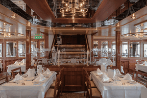 Elegant skibsrestaurant med hvide duge, træpaneler og central trappe. Reflekterende loft og panoramavinduer langs væggene.