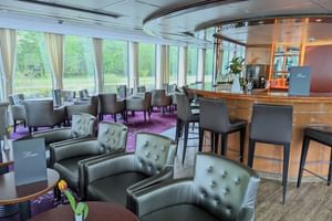 Interiör av MS SE-MANON salong med grå läderfåtöljer, bar med pallar, matbord och stora fönster som visar grönt landskap utanför.