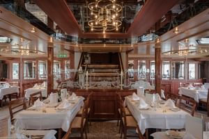 Elegant skibsrestaurant med hvide duge, træstole og historisk skovlhjul som midtpunkt. Spejlloft og varm belysning.