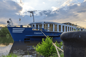 Blåt og hvidt flodkrydstogtskib MS Casanova fortøjet ved en grøn flodbred ved skumring. En pullert med reb er synlig i forgrunden.