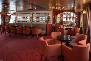 Elegant salon ombord på skibet Switzerland med koralfarvet lænestole, runde borde, rødt tæppe og træpaneleret loft med lysekroner.