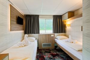 Moderne to-sengs lugare på MS Normandie med to enkeltsenger, trepanel, veggmontert TV, vindu med grå gardiner og mønstret teppe.