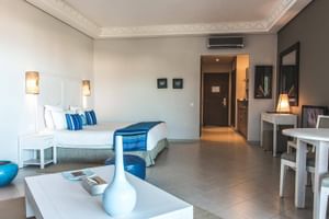 Rummelig junior suite med hvid seng, blå accenter, spisebord og moderne indretning. Flisegulv og dekorative loftslister synlige.