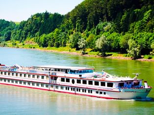 Hvidt flodkrydstogtskib MS Normandie med rød kant sejlende på Donau, omgivet af frodigt grønne skovklædte bakker.