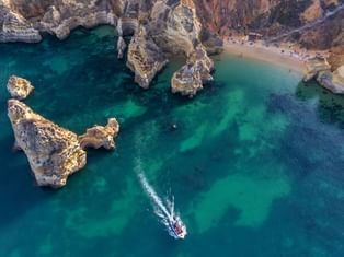 Flyfoto av turkise farvann og gylne klipper i Lagos, Portugal. En liten båt lager et hvitt kjølvann i det smaragdgrønne havet nær fjellformasjoner.