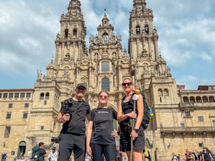 Tre vandrere poserer foran katedralen i Santiago de Compostela på dag 14 av den portugisiske Camino, med barokke tårn bak dem.