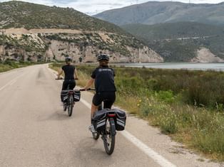 To cyklister med cykeltasker kører på en kystvej i Ithaca, Grækenland. Bjerge og vand synlige i baggrunden med grøn vegetation.