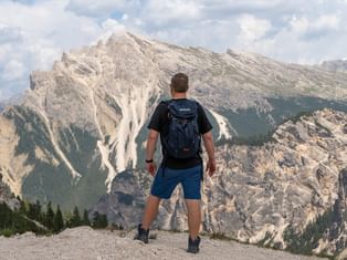 Mannlig fotturist med ryggsekk står på fjellkant og ser på dramatiske kalksteinstopper i Dolomittene under skyet himmel.