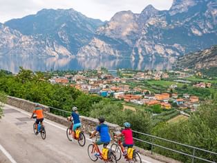 Group of cyclists in colorful jerseys stopped on a mountain road overlooking Lake Garda, with dramatic mountains and a lakeside town below.