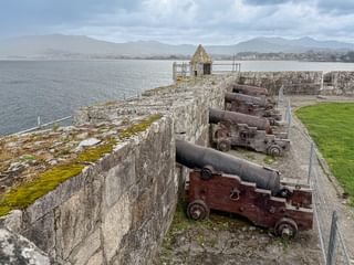 Stenfæstningsvold med historiske kanoner på trælaffetter med udsigt over en bugt. Bjerge synlige over vandet under skyet himmel.