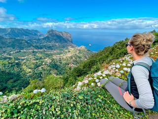 Kvinnelig turgåer med blå sekk sitter blant hvite blomster og ser utover Madeiras dramatiske kyst med fjell og blått hav.