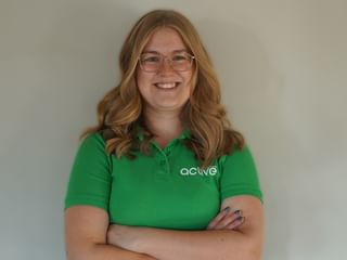 Smilende blond kvinde med briller iført en grøn Active polo, står med korslagte arme mod en hvid væg.