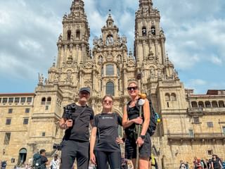 Tre vandrere poserer foran katedralen i Santiago de Compostela på dag 14 av den portugisiske Camino, med barokke tårn bak dem.
