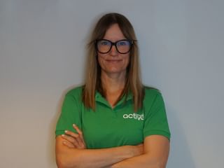 Professionelt portræt af Edith med briller og grøn poloshirt med 'active' logo, stående med korslagte arme foran hvid væg.