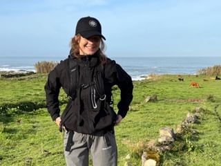 Kvinde ved navn Katrine med sort kasket og jakke på grøn kysteng med hav og græssende dyr i baggrunden.