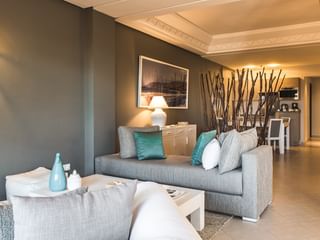 Moderne stue med grå sofa, turkise puder, hvidt sofabord og dekorativ grenskærm. Varm belysning og beige flisegulv.