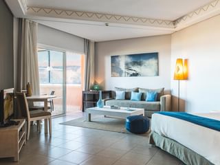 Rummelig junior suite med seng, sofa, spiseplads og adgang til balkon. Blå og beige indretning med varm belysning og dekorativ vægkunst.