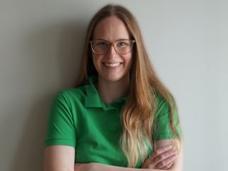 Portræt af Emelia med briller og grøn poloshirt, smilende med korslagte arme mod en hvid vægbaggrund.