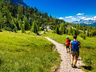 Two hikers walking on a gravel mountain path through green meadows in the Dolomites, with snow-capped peaks and dense forests visible.