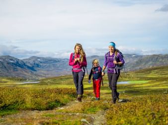Tre vandrere på Kongevegen gennem grønt bjerglandskab i Norge med bjerge og sø i baggrunden.