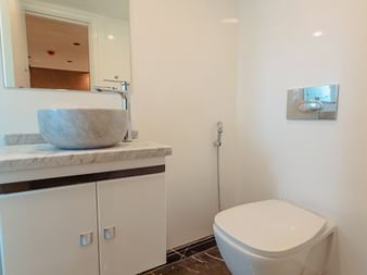 Moderne badeværelse med hvid marmor bordplade, rund påbygget håndvask, vægmonteret toilet og hvide skabe. Spejlet reflekterer tilstødende rum.