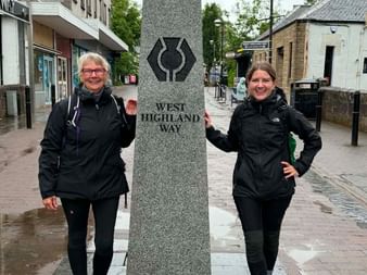 To kvinder i sorte regnjakker står ved en granitmarkør for West Highland Way på en skotsk byplads på en regnfuld dag.