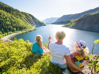 Tre personer sidder på en græsklædt skråning med udsigt over en blå fjord omgivet af grønne skovklædte bjerge langs Kongevegen i Norge.