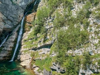 Vattenfall som faller ner över klippiga branter till en turkos pool i Juliska Alperna. Branta kalkstenväggar täckta med grön vegetation ramar in scenen.