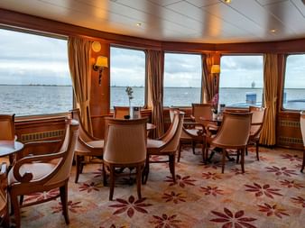 Elegant salon ombord på MS Rigoletto med buede vinduer ud mod vandet, med træstole omkring borde og blomstret tæppe.